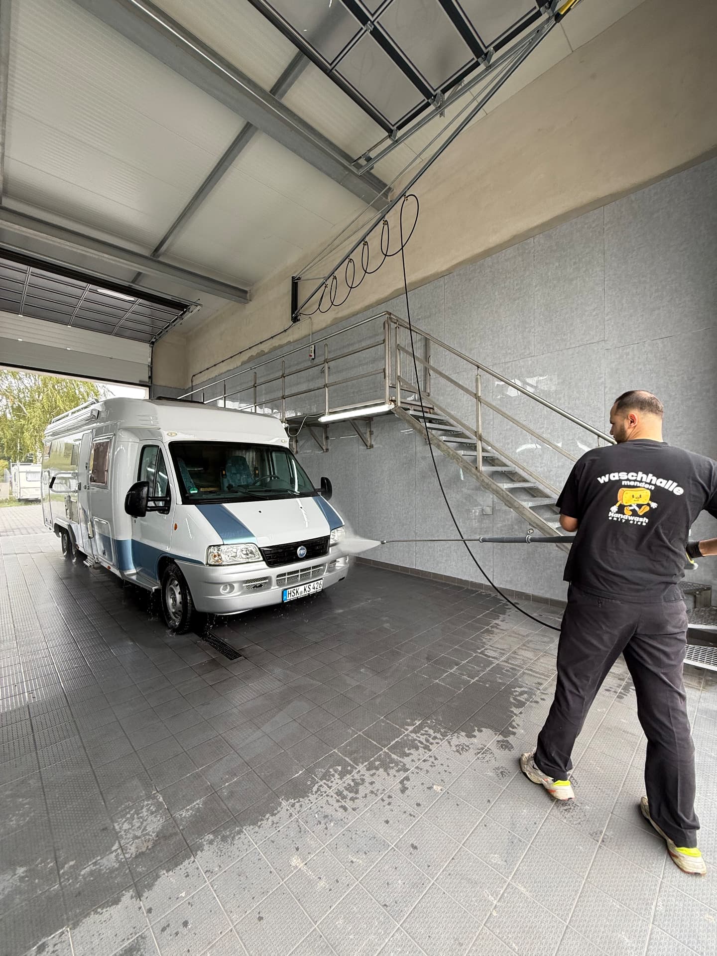 Team der Waschhalle wäscht die Front eines Wohnmobils mit der Hochdrucklanze neben der Treppe zur Empore